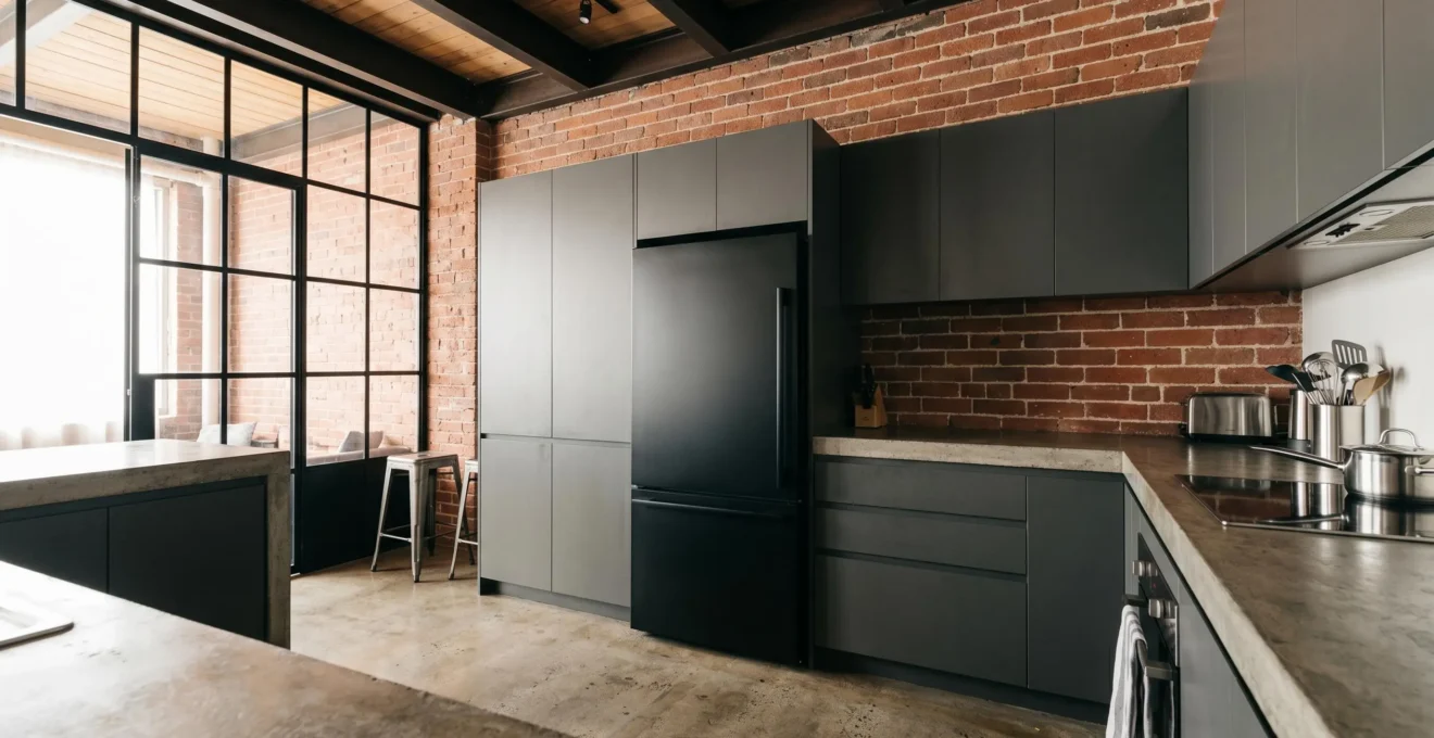 Cuisine de loft au style industriel avec murs en briques apparentes, poutrelles métalliques et réfrigérateur encastrable noir mat intégré dans la rangée de meubles